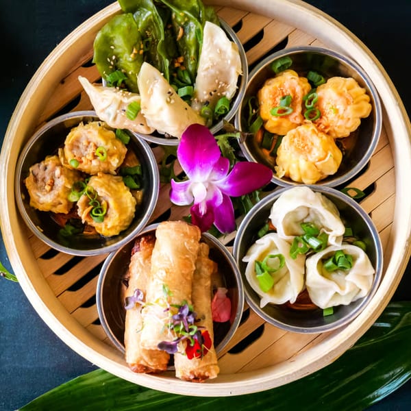 A bamboo steamer basket filled with various dim sum, including dumplings, spring rolls, and a purple orchid.
