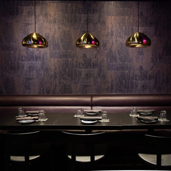 Dark, modern dining area with three tables set with glasses and napkins, under golden pendant lights.