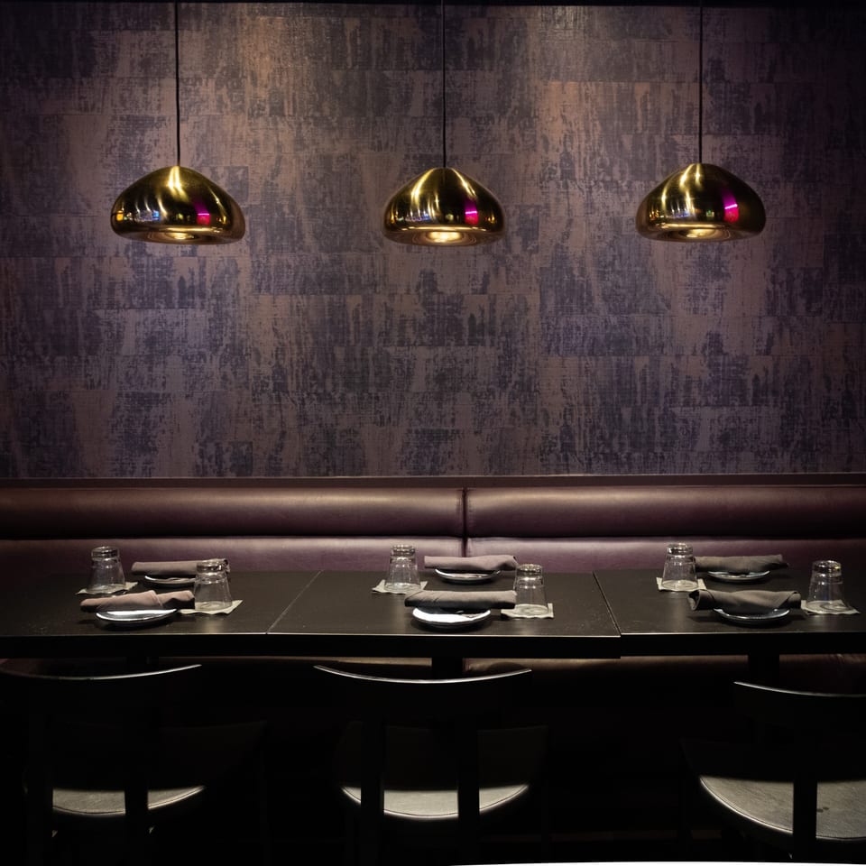 Dark, modern dining area with three tables set with glasses and napkins, under golden pendant lights.