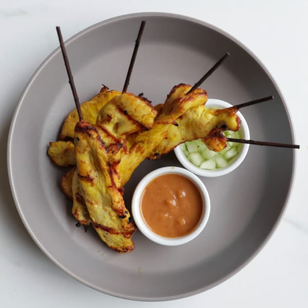 Grilled chicken satay skewers with peanut sauce and cucumber relish on a gray plate.