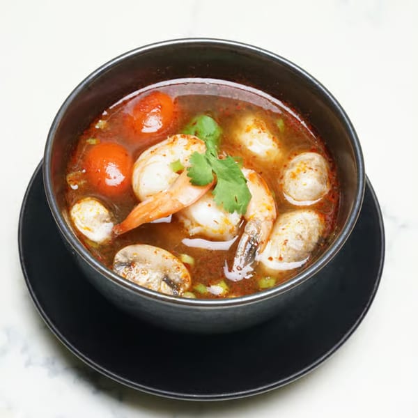 A black bowl of Tom Yum soup with shrimp, mushrooms, and cherry tomatoes.