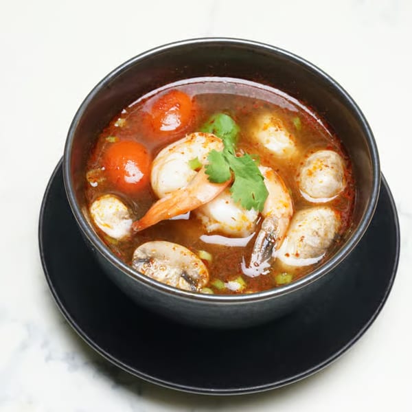 A black bowl of Tom Yum soup with shrimp, mushrooms, and cherry tomatoes, garnished with cilantro.