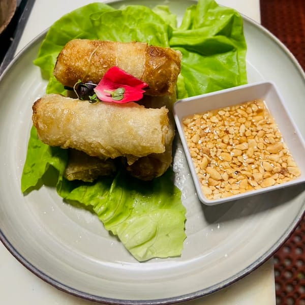 Crispy spring rolls on lettuce with a side of peanut dipping sauce, garnished with a pink flower.