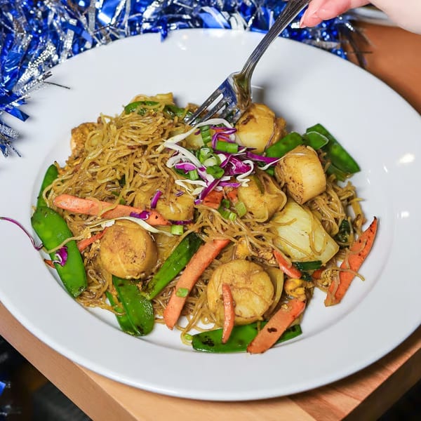 A fork lifts noodles and scallops from a plate of stir-fried noodles with vegetables.
