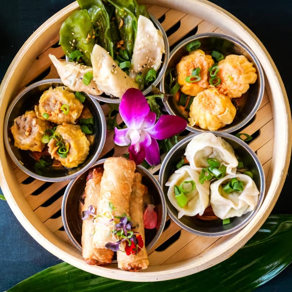 A bamboo steamer basket filled with various types of dumplings, spring rolls, and a purple orchid.