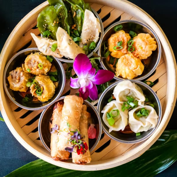 A bamboo steamer basket filled with various dim sum, including dumplings, spring rolls, and a purple orchid.