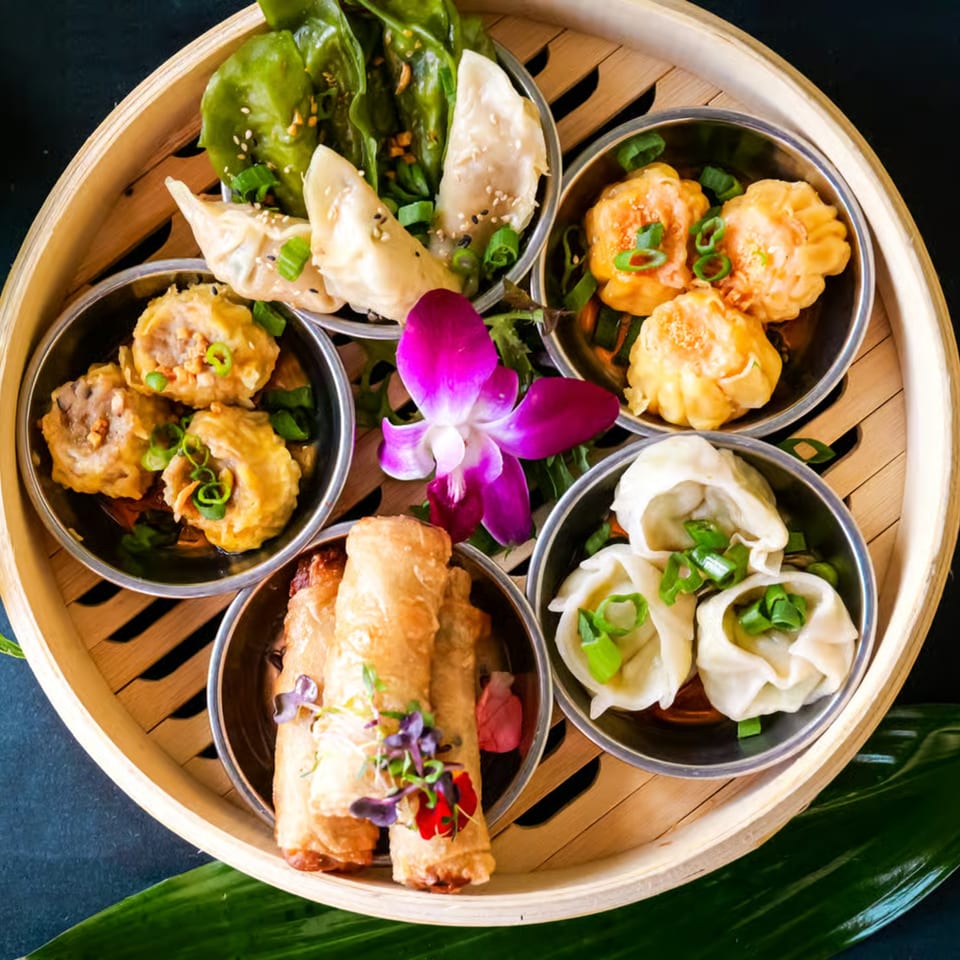 A bamboo steamer basket filled with various dim sum, including dumplings, spring rolls, and a purple orchid.